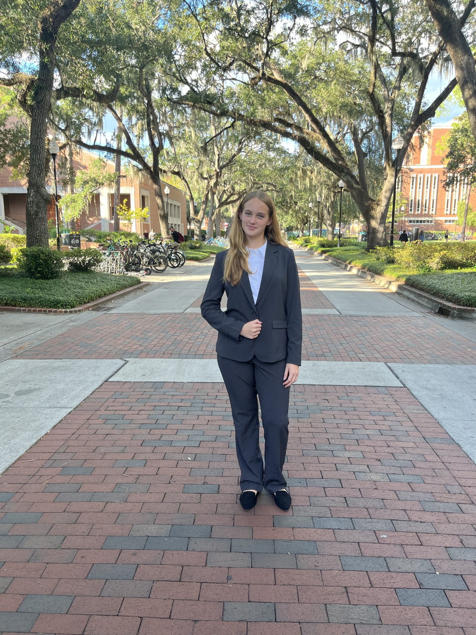 Image of young woman with long blond hair in a grey pant suit standing on a walking path covered in trees. 